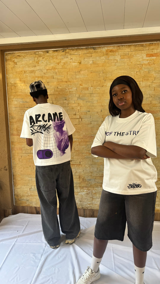Two models wearing white graphic t-shirts in an indoor setting with a brick wall background. The model on the left is shown from the back wearing a white t-shirt with &